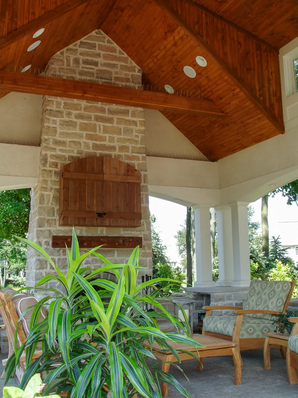 Covered pavilion interior with stone fireplace, warm wood ceiling, seating, columns, and potted plant.